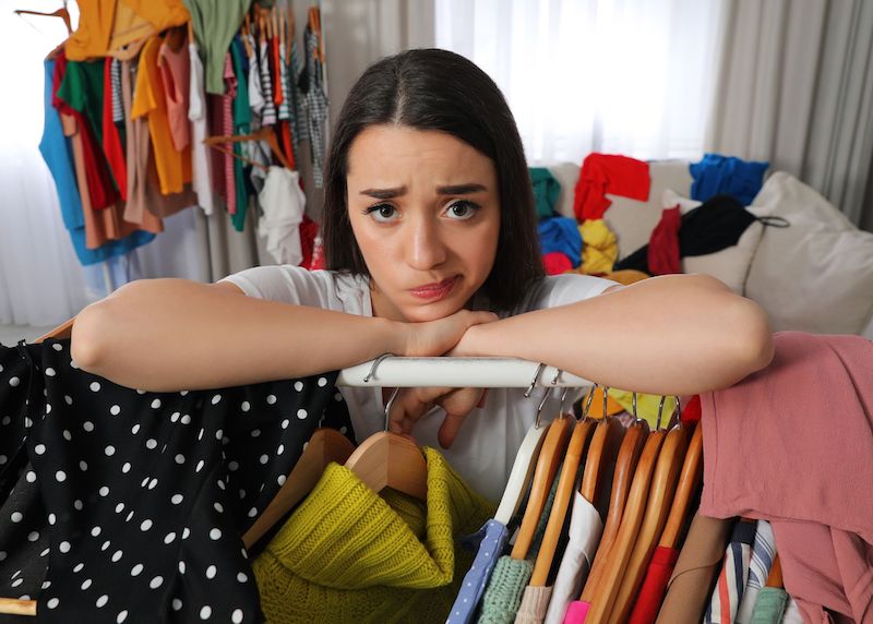 puzzled woman doing a closet edit leans on clothes rack surrounded by clothes